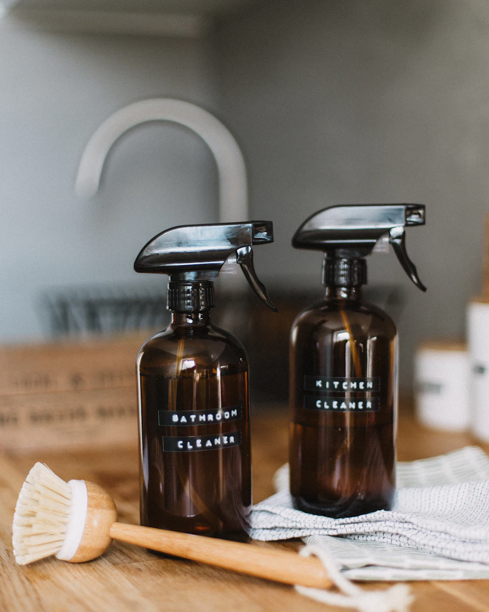 Two amber glass spray bottles and a wooden dish brush on a linen cloth