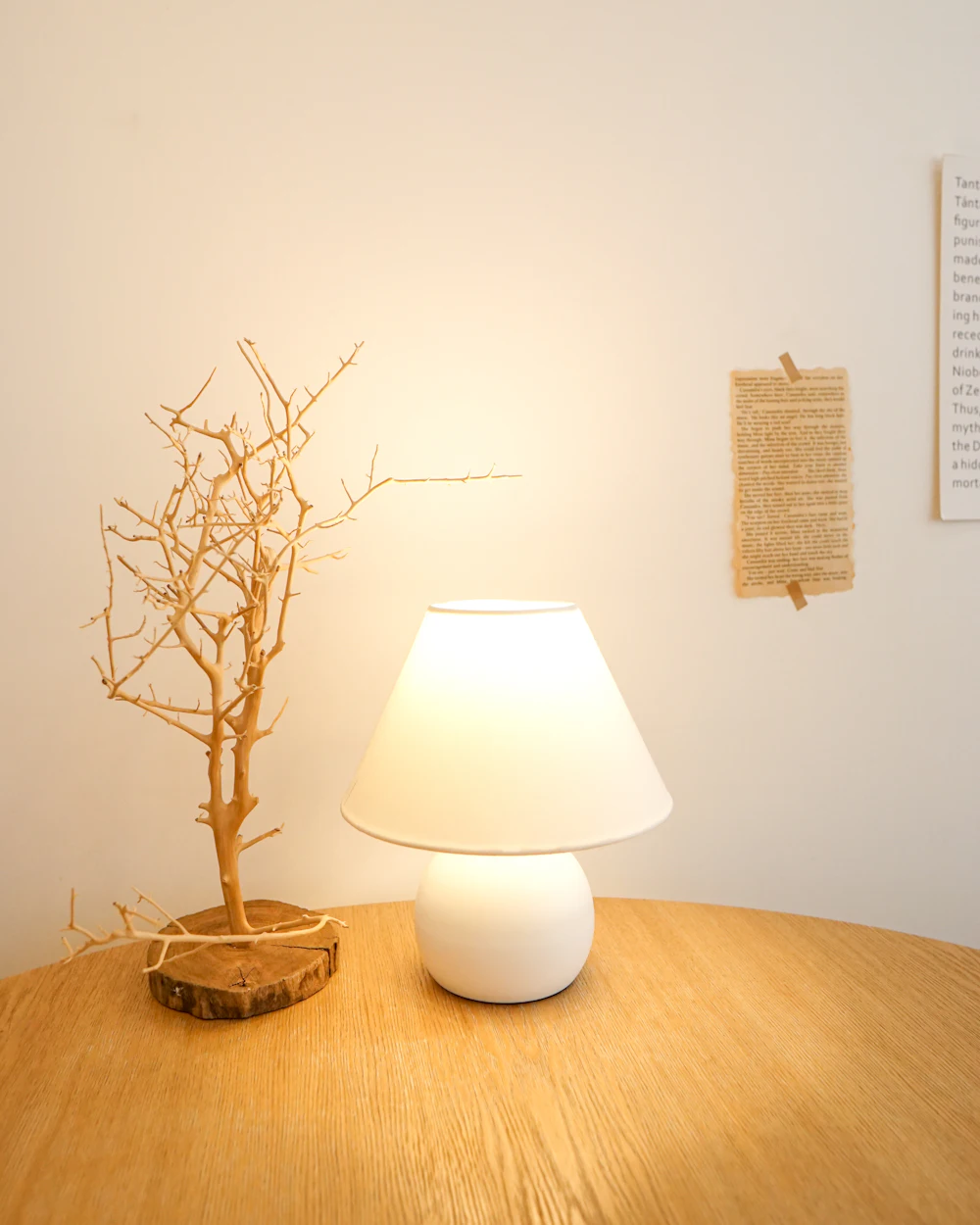 A small white table lamp glowing next to a dried branch on a round wooden table