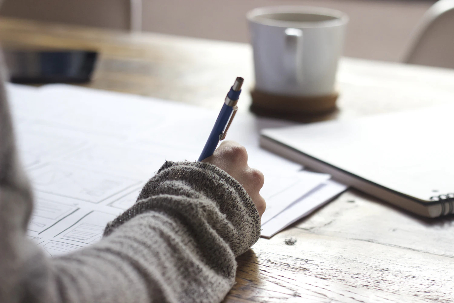 A hand writing notes in a notebook beside a ceramic mug on a wooden table