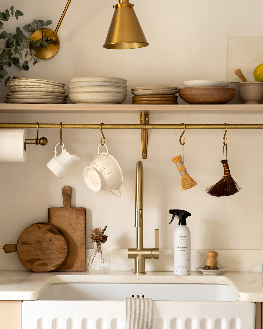 A sunlit kitchen corner with a brass wall lamp, an open shelf of bowls and a white farmhouse sink