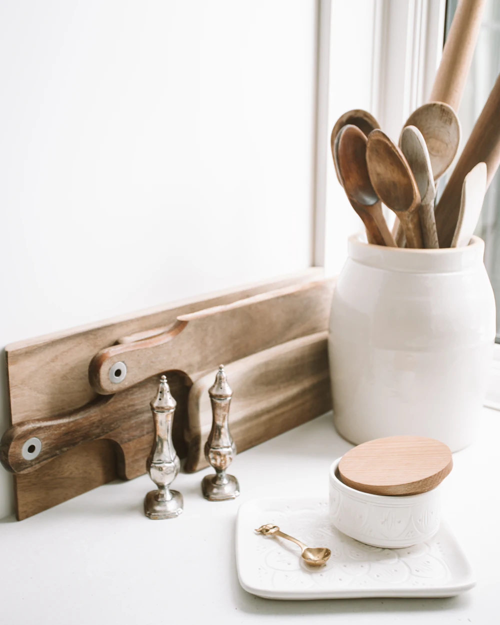 Wooden spoons in a white ceramic crock beside a cutting board on a bright counter