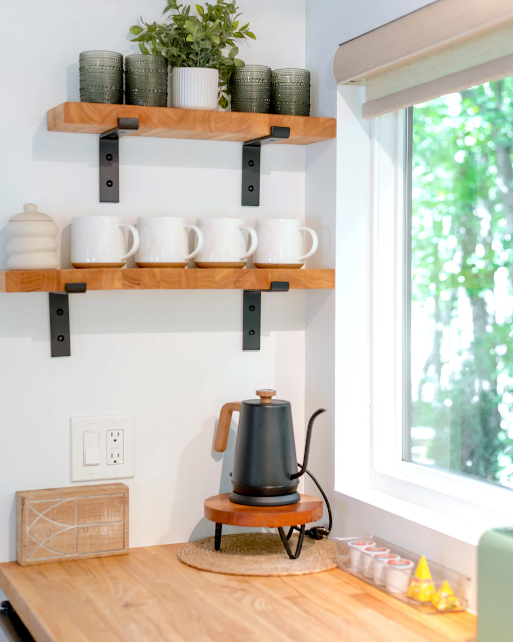 Wooden shelves with white mugs above a butcher-block counter and a black kettle by a window